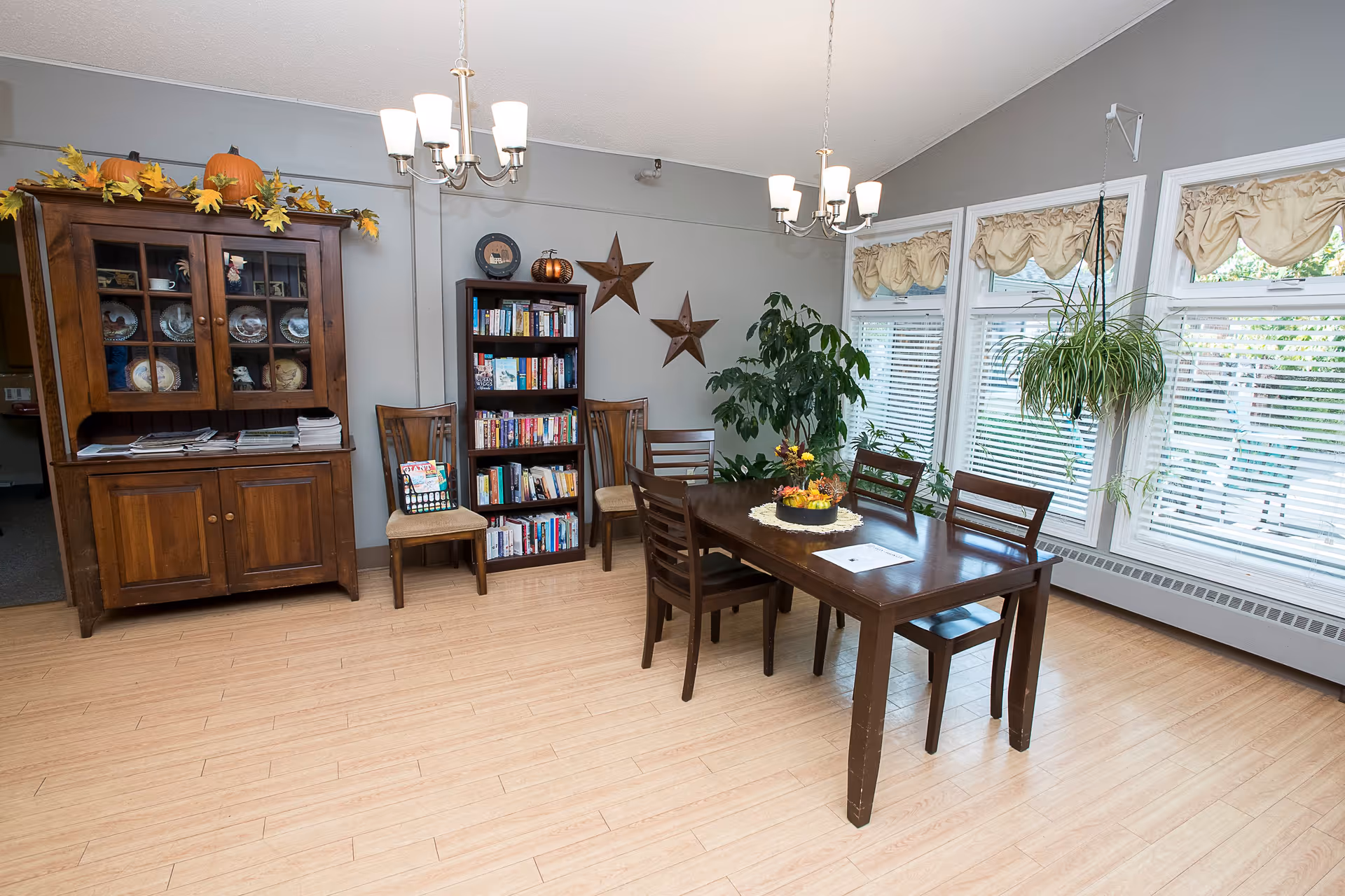 A well-lit dining area with a wooden dining table surrounded by six chairs. The room features large windows with beige valances, a wooden cabinet with decorative plates and pumpkins on top, a bookshelf filled with books, two star-shaped wall decorations, and several green plants including a hanging plant near the windows. The floor is light wood, and two chandeliers hang from the ceiling.