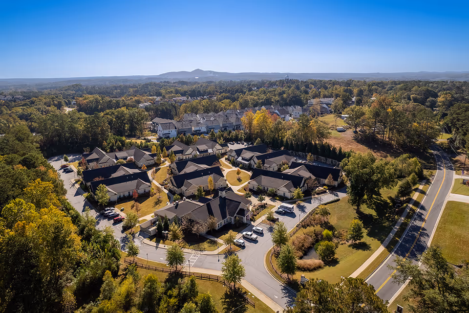 Aerial view of Camellia Place an Oaks Senior Living Community showing multiple single-story buildings with dark roofs arranged in a circular layout surrounded by trees and greenery, with a road curving around the community and a clear blue sky above.