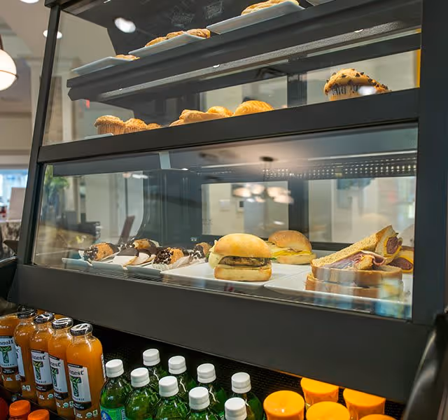 A glass display case showing sandwiches and pastries with bottled drinks below in a dining area.
