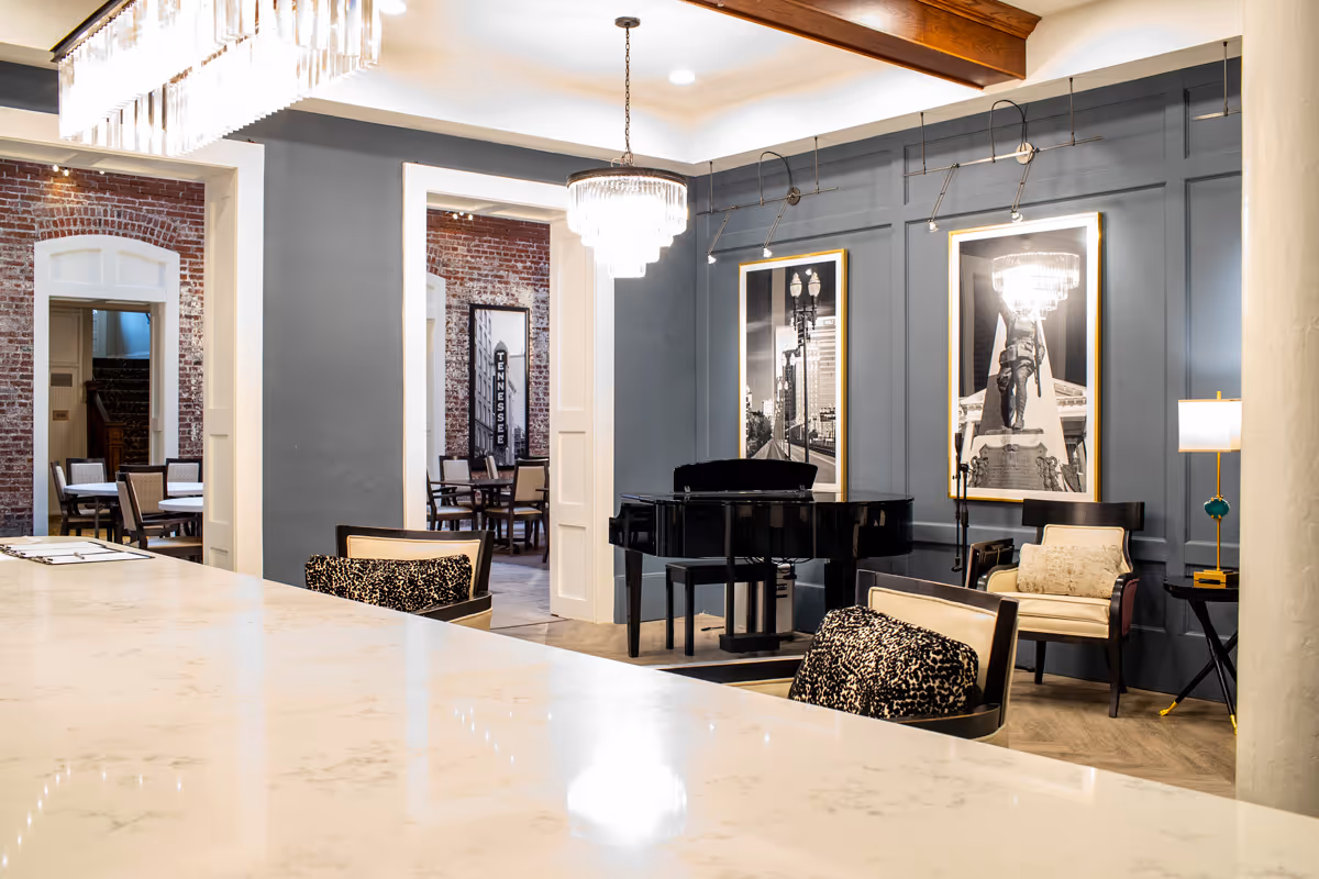 Interior view of a stylish common area featuring a black grand piano, two armchairs with patterned cushions, framed black and white cityscape photographs on a blue paneled wall, a modern chandelier, and a marble countertop in the foreground. Adjacent rooms with exposed brick walls and dining tables with chairs are visible through open doorways.