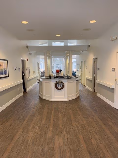 Interior hallway of a senior living facility with wood flooring, white walls, and a central reception desk decorated with a wreath. The hallway has multiple doorways on either side and recessed ceiling lights.