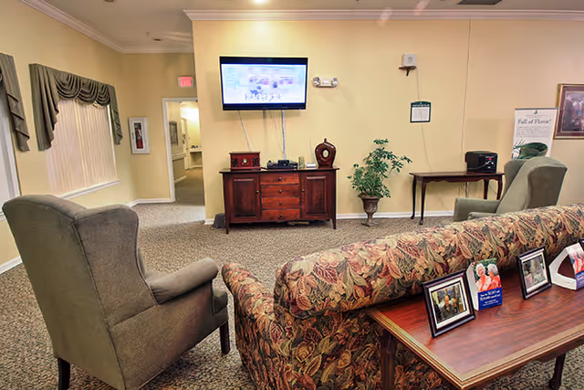A cozy living room area in a senior living facility with a floral patterned sofa and a gray armchair facing a wall-mounted television. A wooden console with decorative items is beneath the TV, and a small table with a plant and another piece of furniture are against the wall. The room has beige walls, carpeted floor, and a window with draped curtains.
