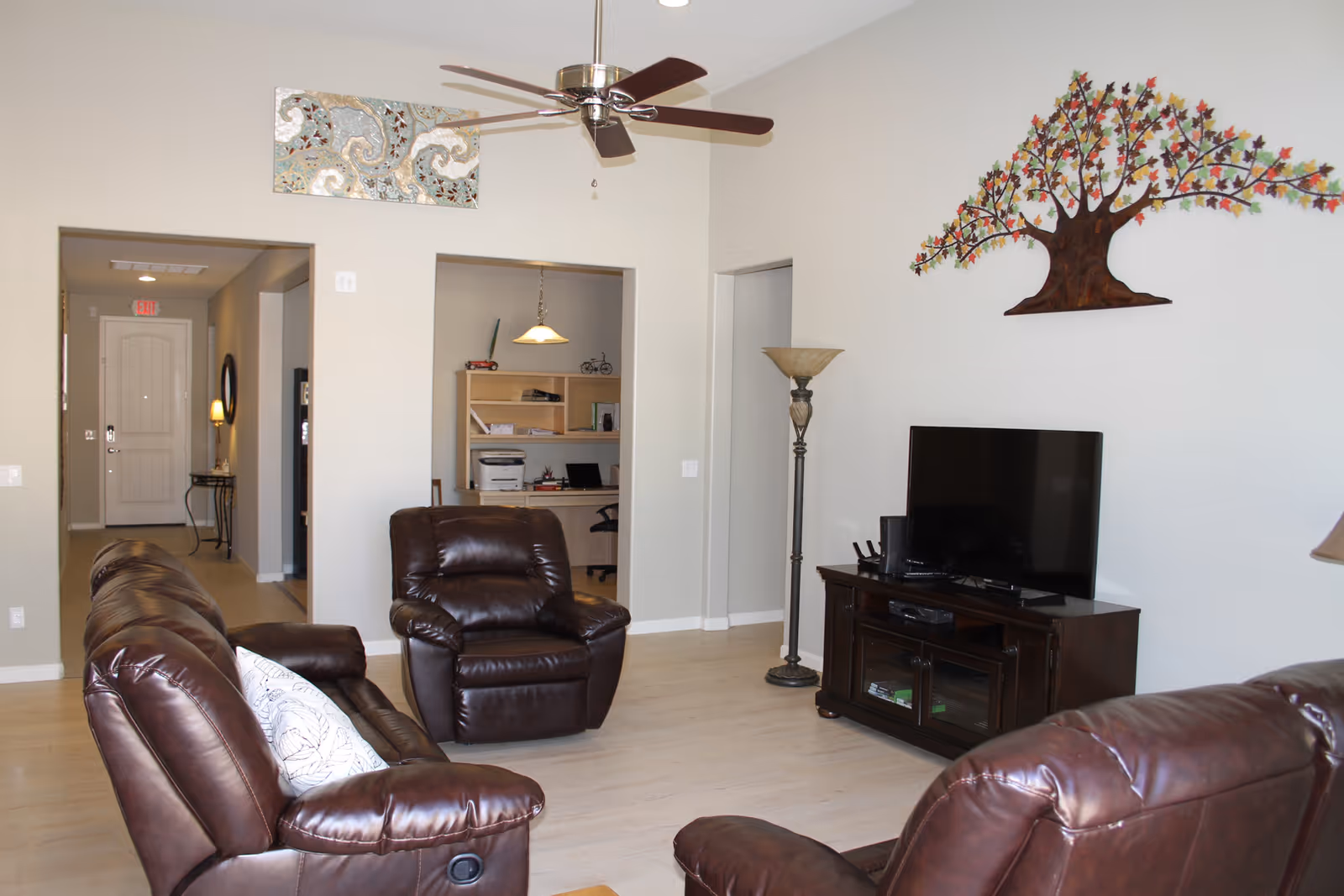 A living room with brown leather recliner chairs and a matching sofa arranged around a dark wooden TV stand with a flat-screen television. The walls are light-colored with a decorative metal tree wall art above the TV and a ceiling fan with wooden blades. In the background, there is a hallway leading to a door and an office area with a desk, chair, and shelves.