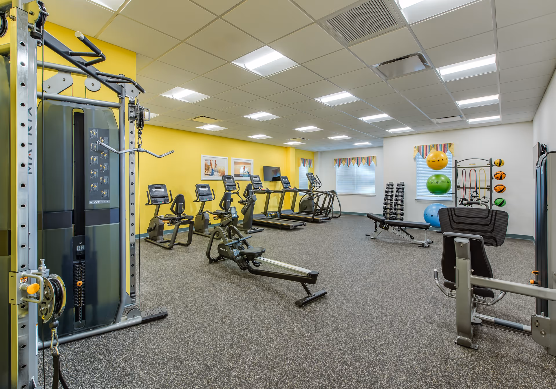 A bright fitness room with various exercise equipment including treadmills, stationary bikes, a rowing machine, weight bench, dumbbells, and colorful exercise balls. The walls are painted yellow and white, with two windows featuring striped valances. The room has a carpeted floor and a drop ceiling with fluorescent lighting.