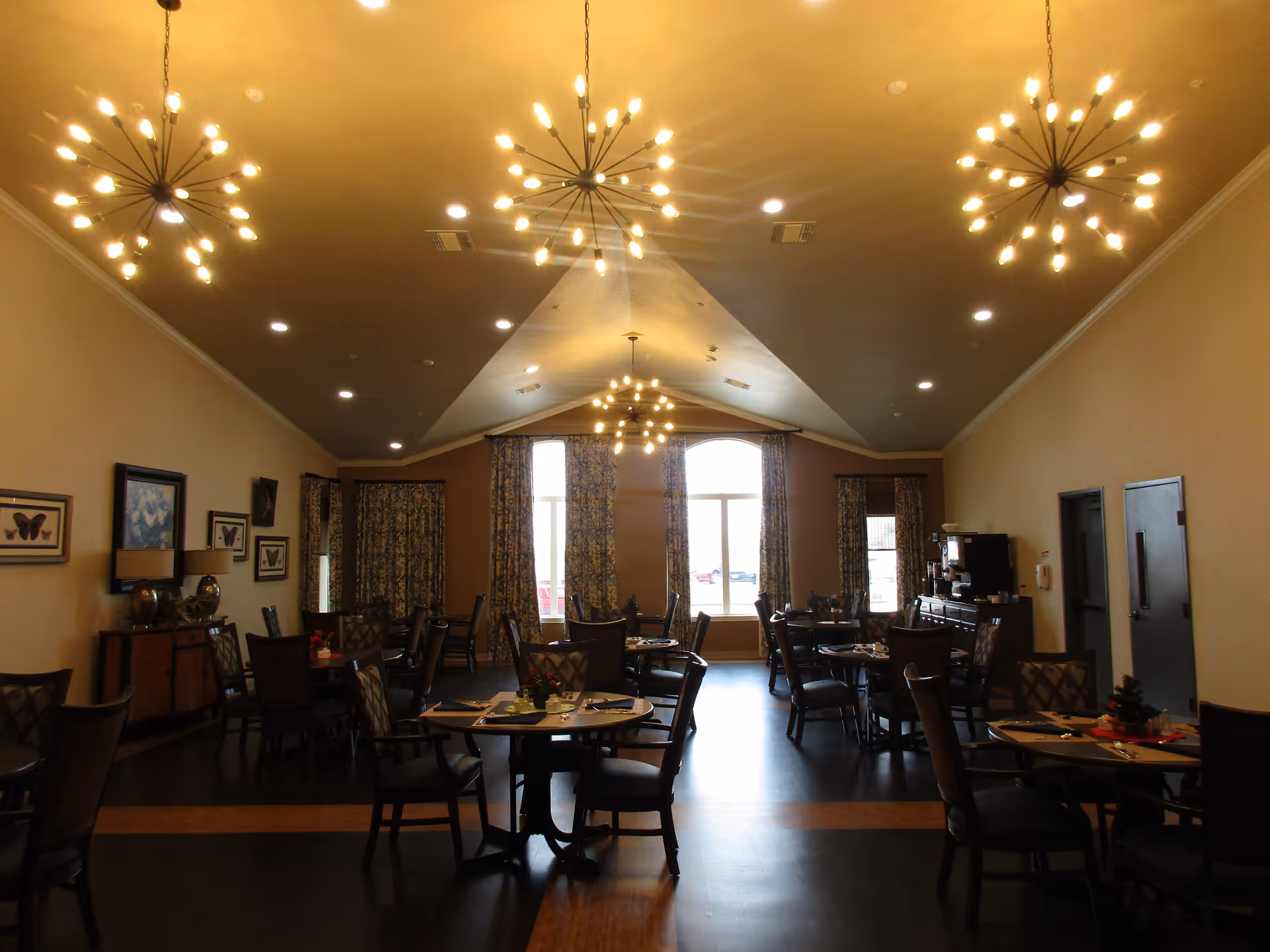 Spacious assisted-living dining room with round tables and chairs, patterned curtains, and starburst chandeliers.