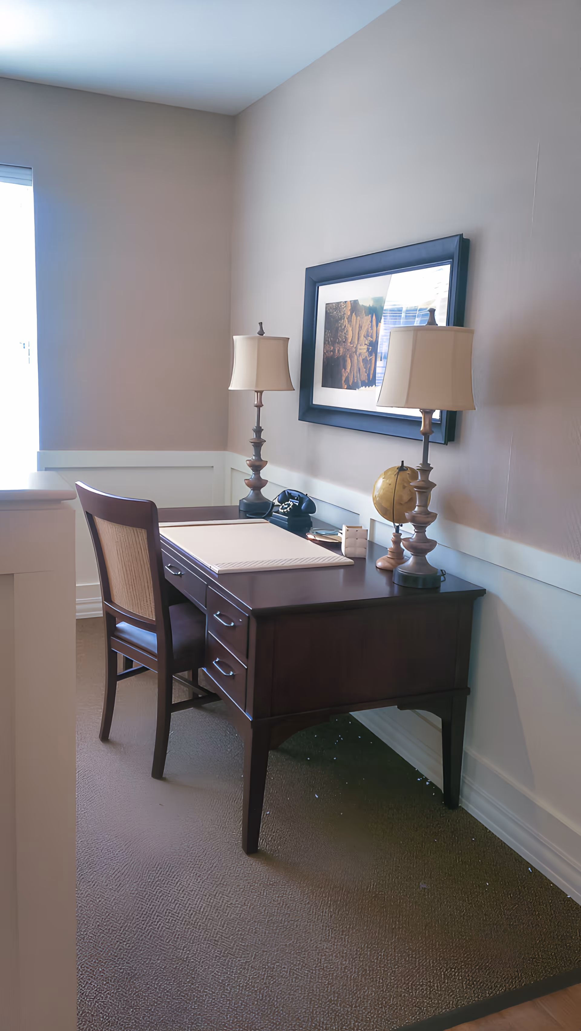 A small office area with a dark wooden desk and matching chair. The desk has two table lamps with beige shades, a black rotary phone, a small globe, and a framed picture hanging on the wall above it. The room has beige walls and carpeted floor.