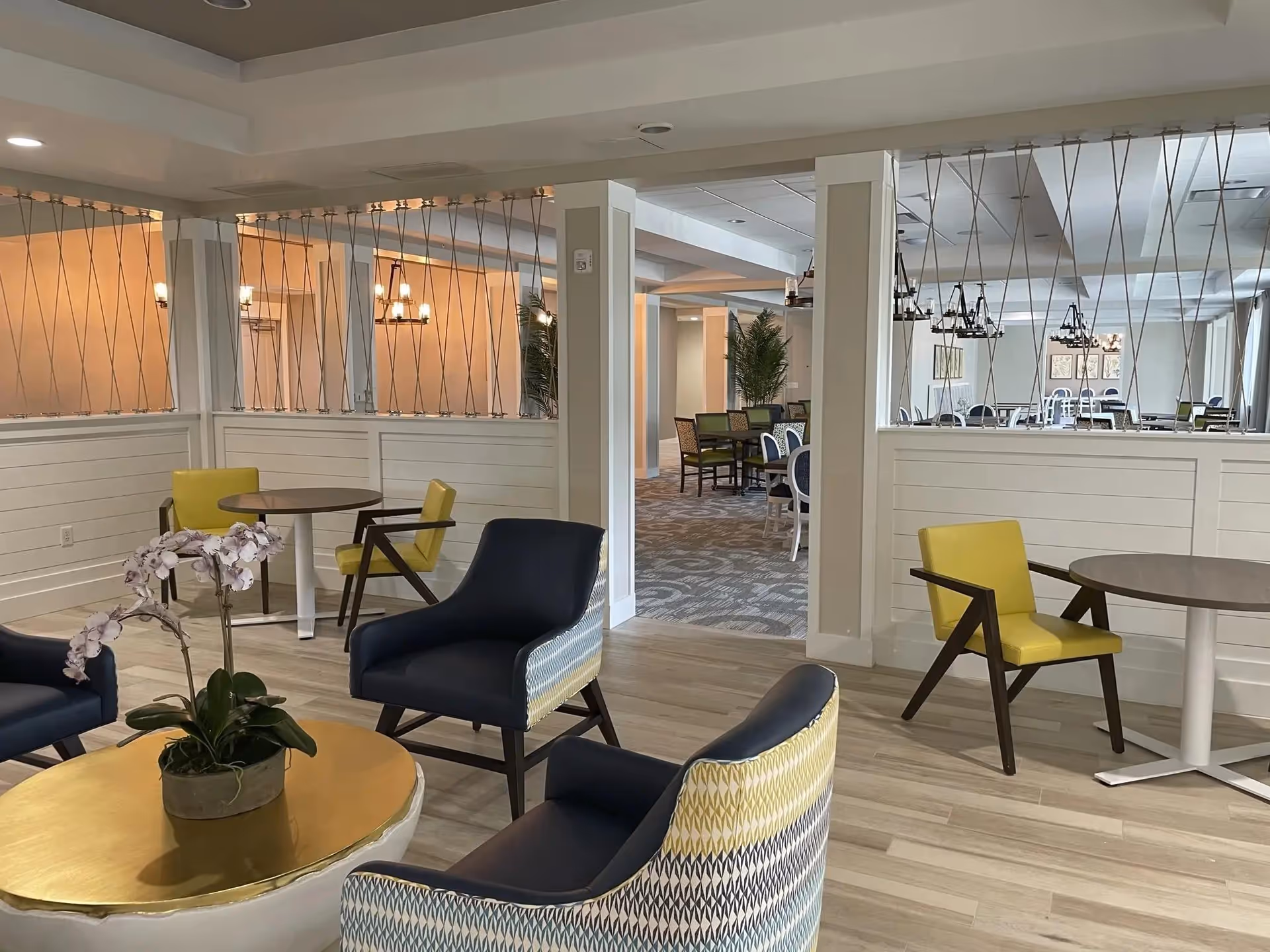 A modern and spacious common area in a senior living facility featuring comfortable seating with blue and yellow chairs around small round tables. The room has light wood flooring, white paneled half-walls with decorative metal rods, and warm lighting. In the background, there is a dining area with multiple tables and chairs, and some greenery for decoration.