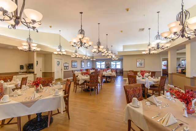 A spacious dining room in a senior living facility with multiple tables covered in white tablecloths, set with plates, cups, napkins, and silverware. Each table has a centerpiece with red flowers. The room features wooden chairs with patterned upholstery, warm lighting from chandeliers hanging from the ceiling, and windows along the far wall letting in natural light.