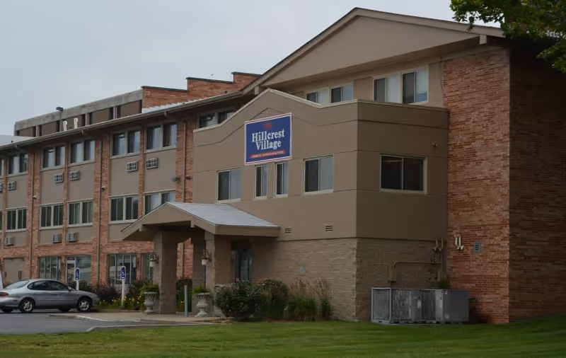 Front exterior of the multi-story Hillcrest Village senior living building with an entrance canopy, sign, parked cars, and lawn.