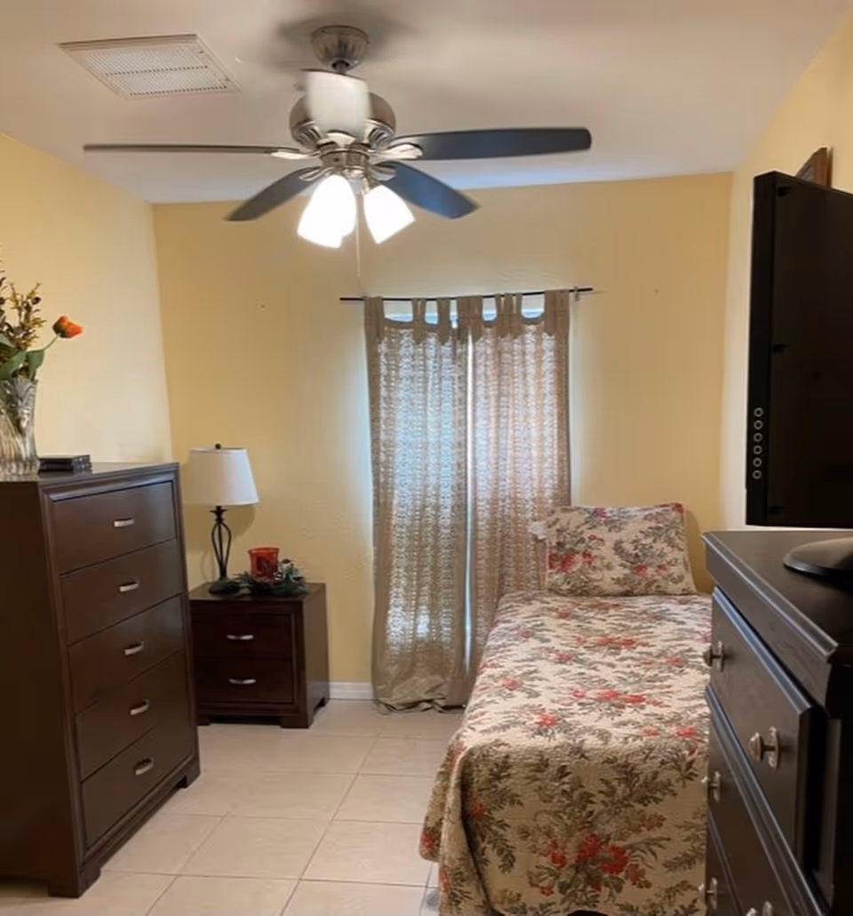 Small bedroom with a single bed in floral bedding, wooden dressers, a nightstand with a lamp, a ceiling fan, and a curtained window.