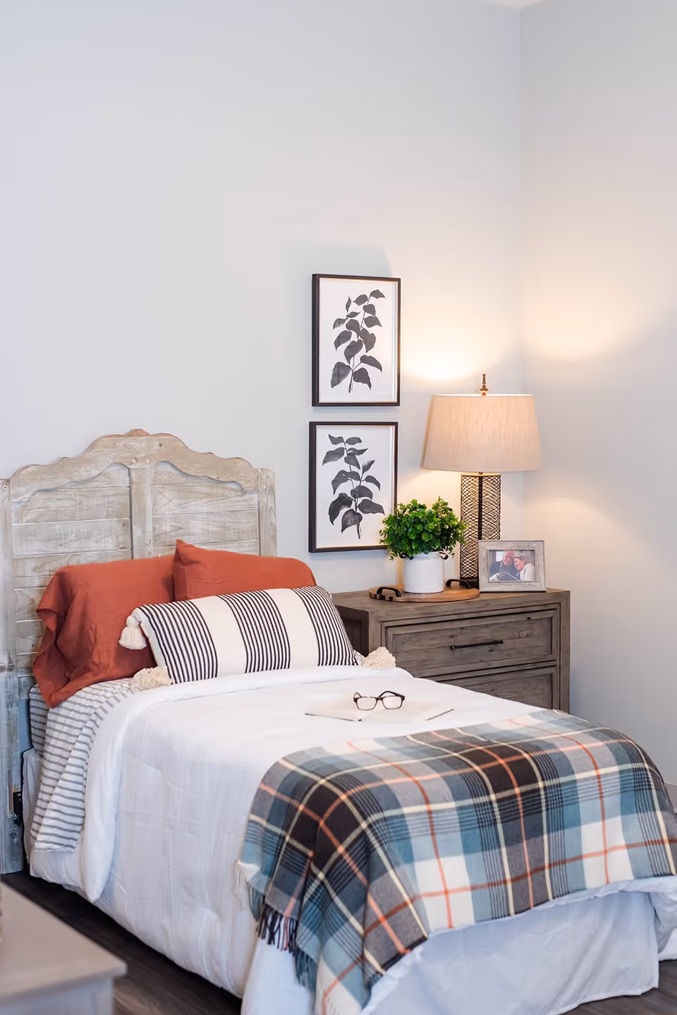 A cozy bedroom with a single bed featuring a rustic wooden headboard, white bedding, a striped bolster pillow, and a plaid blanket. Next to the bed is a wooden nightstand with a table lamp, a small green plant, and a framed photo. Two framed botanical prints hang on the wall above the nightstand.