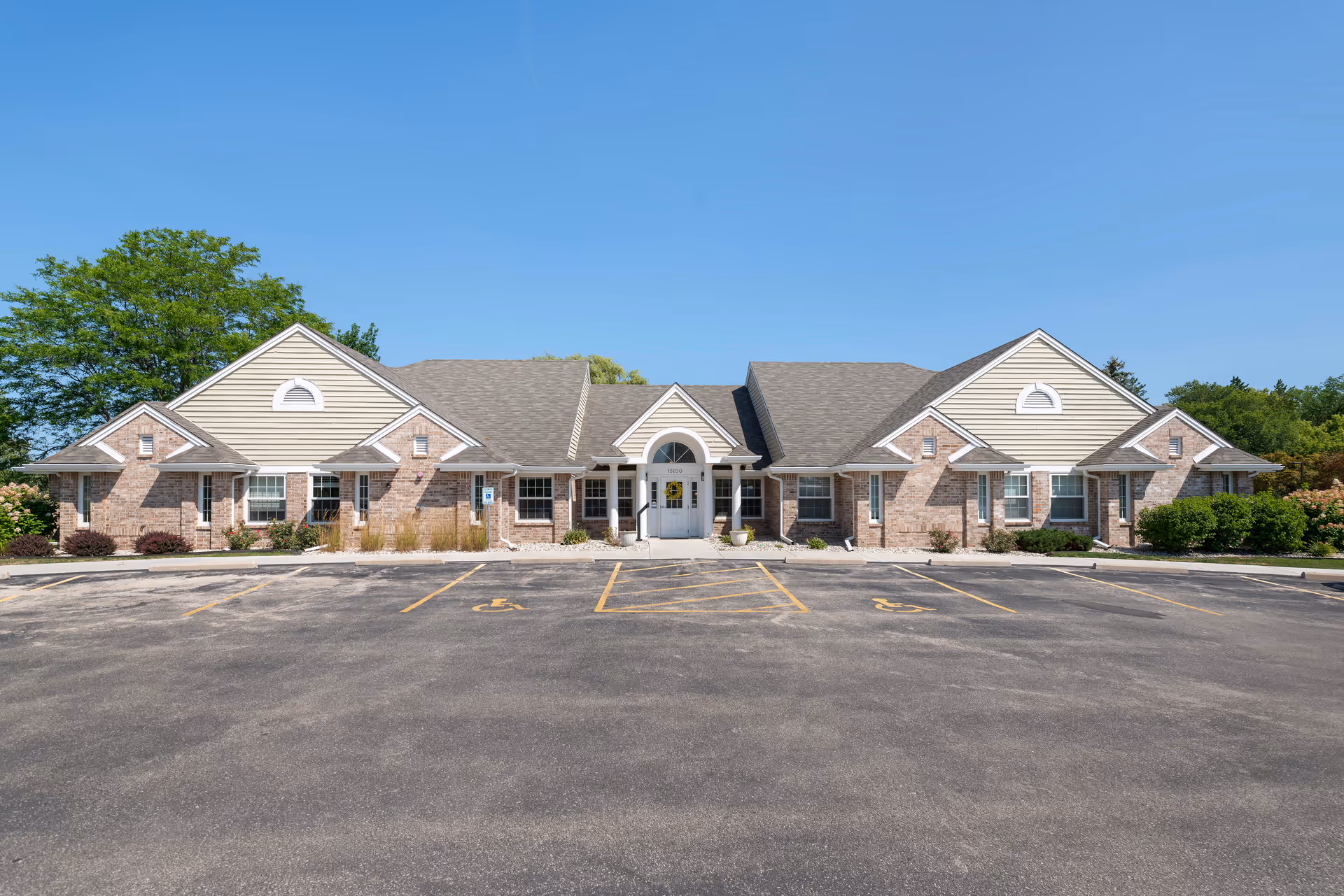 Front exterior view of a single-story senior living facility building with a symmetrical design, brick and beige siding, multiple windows, and a central entrance under a small portico. The building is surrounded by greenery and has a parking lot with marked handicap spaces in front.