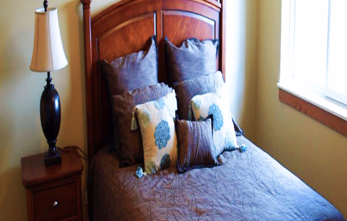 A neatly made bed with decorative pillows and a wooden headboard, beside a nightstand with a lamp and a window.