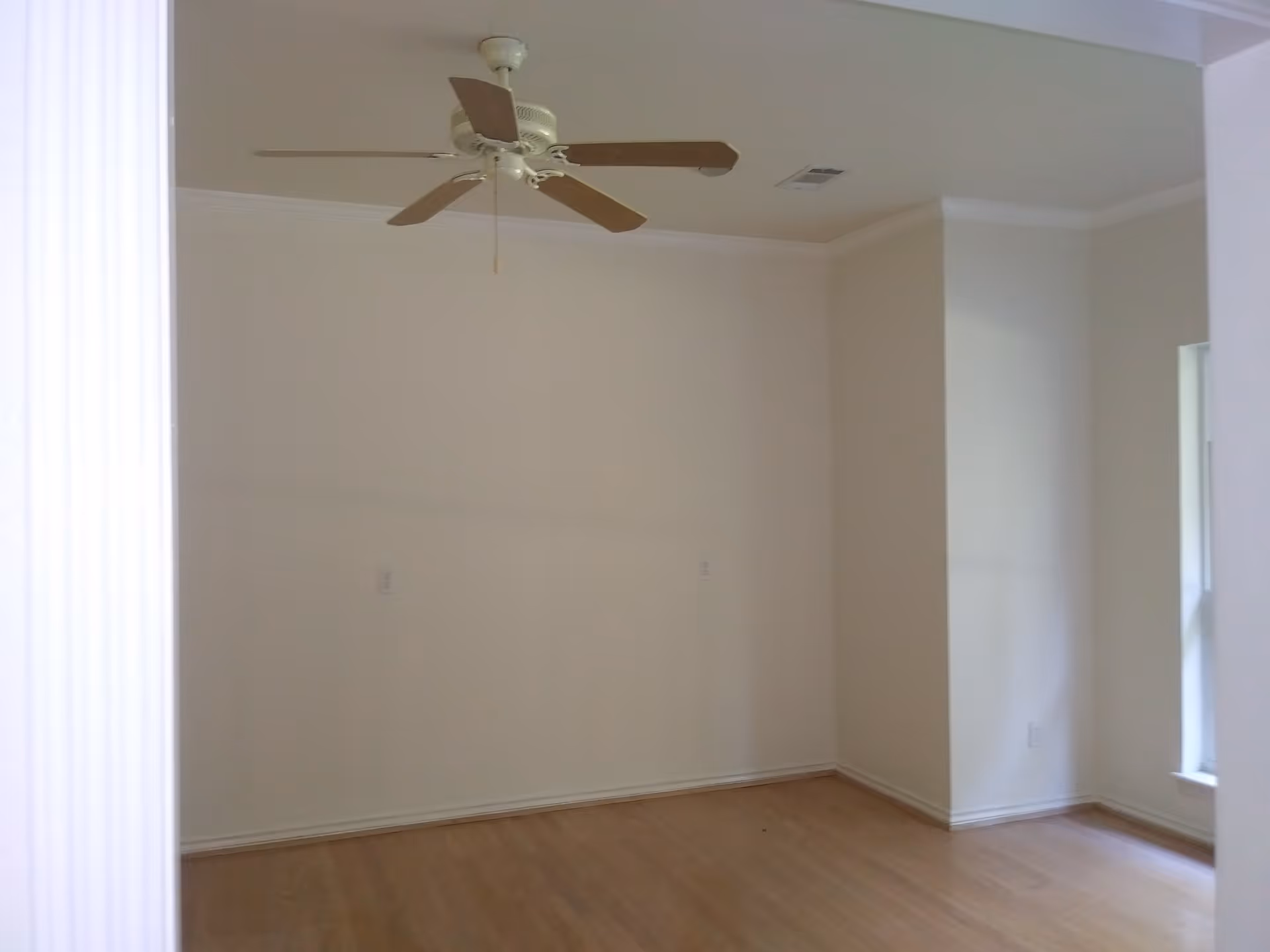 Empty room with light-colored walls, wooden floor, a ceiling fan with wooden blades, and a window on the right side letting in natural light.