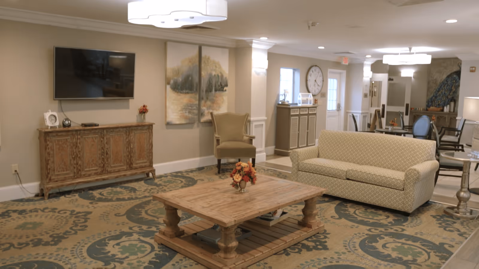 A cozy living room area in The Retreat at Summerville featuring a patterned beige sofa, a wooden coffee table with a floral centerpiece, an armchair, a large wall-mounted TV above a rustic wooden cabinet, and a decorative carpet with intricate designs. The room is softly lit with ceiling lights and wall sconces, and there are paintings and a large clock on the walls. In the background, there are dining tables and chairs, and a door leading outside.