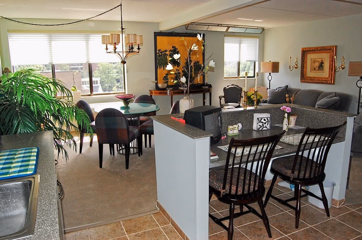 Interior view of a senior living apartment showing a kitchen counter with two wooden chairs, a small TV, and decorative items. Beyond the counter is a living room area with a gray sofa, armchair, lamps, and framed artwork on the wall. To the left is a dining area with a round glass table and four chairs, a chandelier above, and large windows letting in natural light. A large green plant is visible near the kitchen sink.
