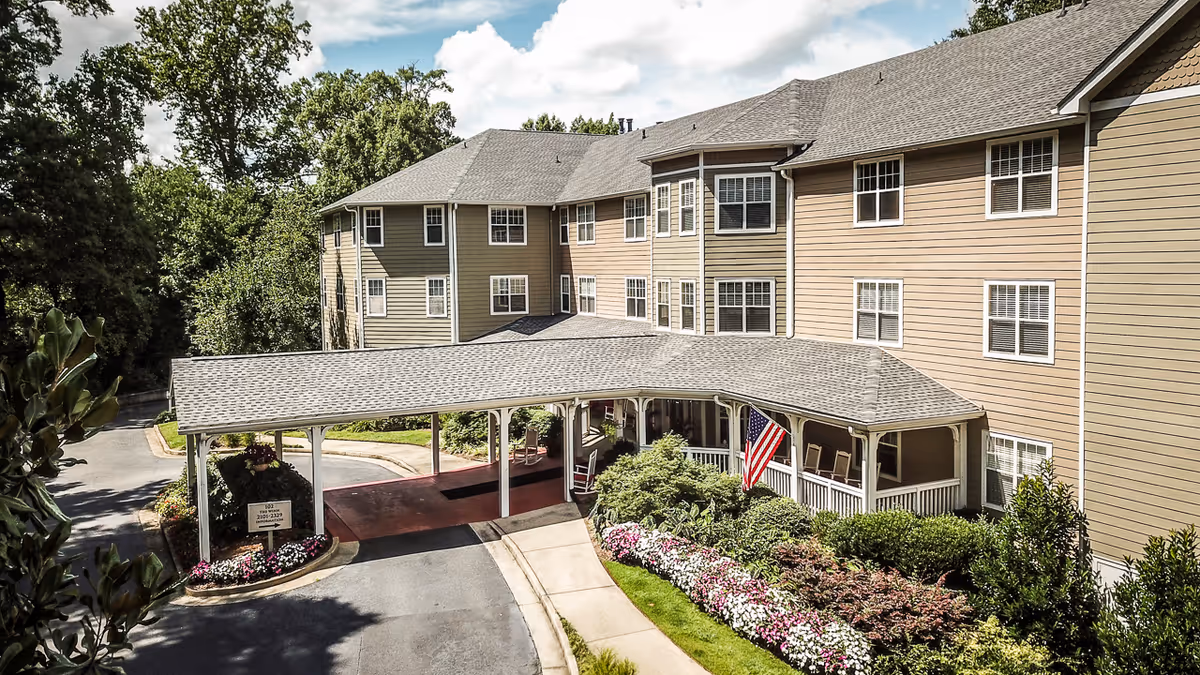 Exterior front of a multi-story senior living building with a covered porte-cochere, American flag, and landscaped flower beds.