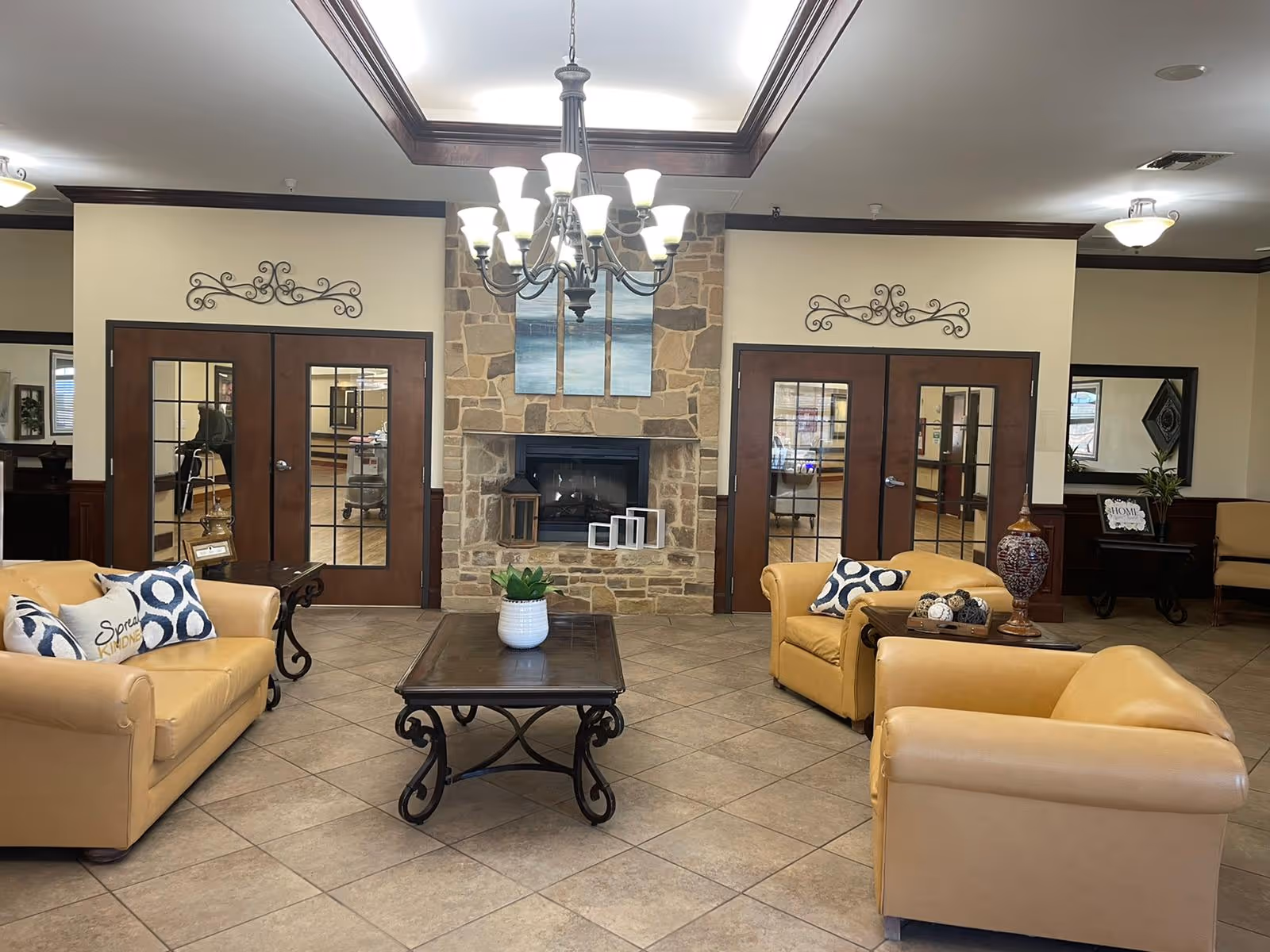 A cozy living room area with tan leather sofas and armchairs arranged around a dark wooden coffee table with a potted plant. The room features a stone fireplace with decorative lanterns and artwork above it. There are double glass doors on either side of the fireplace, and a chandelier hangs from the ceiling. The floor is tiled, and the walls are painted in neutral tones with dark wood trim.