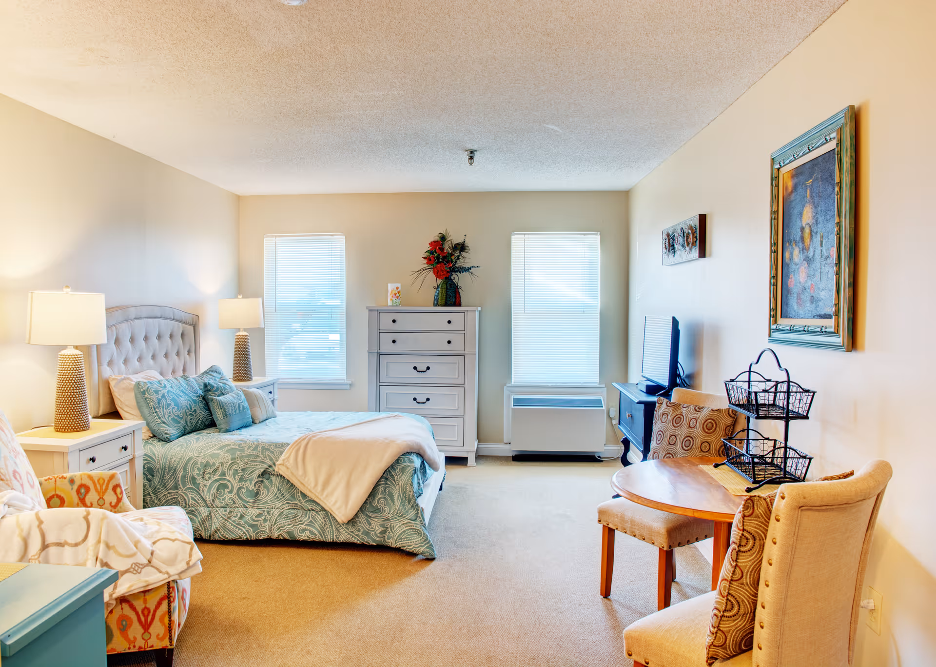 A bright and cozy bedroom with a bed featuring a tufted headboard and teal patterned bedding. There are two bedside tables with lamps, a chest of drawers with a floral arrangement, two windows with blinds, a TV on a stand, a small round wooden table with two upholstered chairs, and framed artwork on the walls.