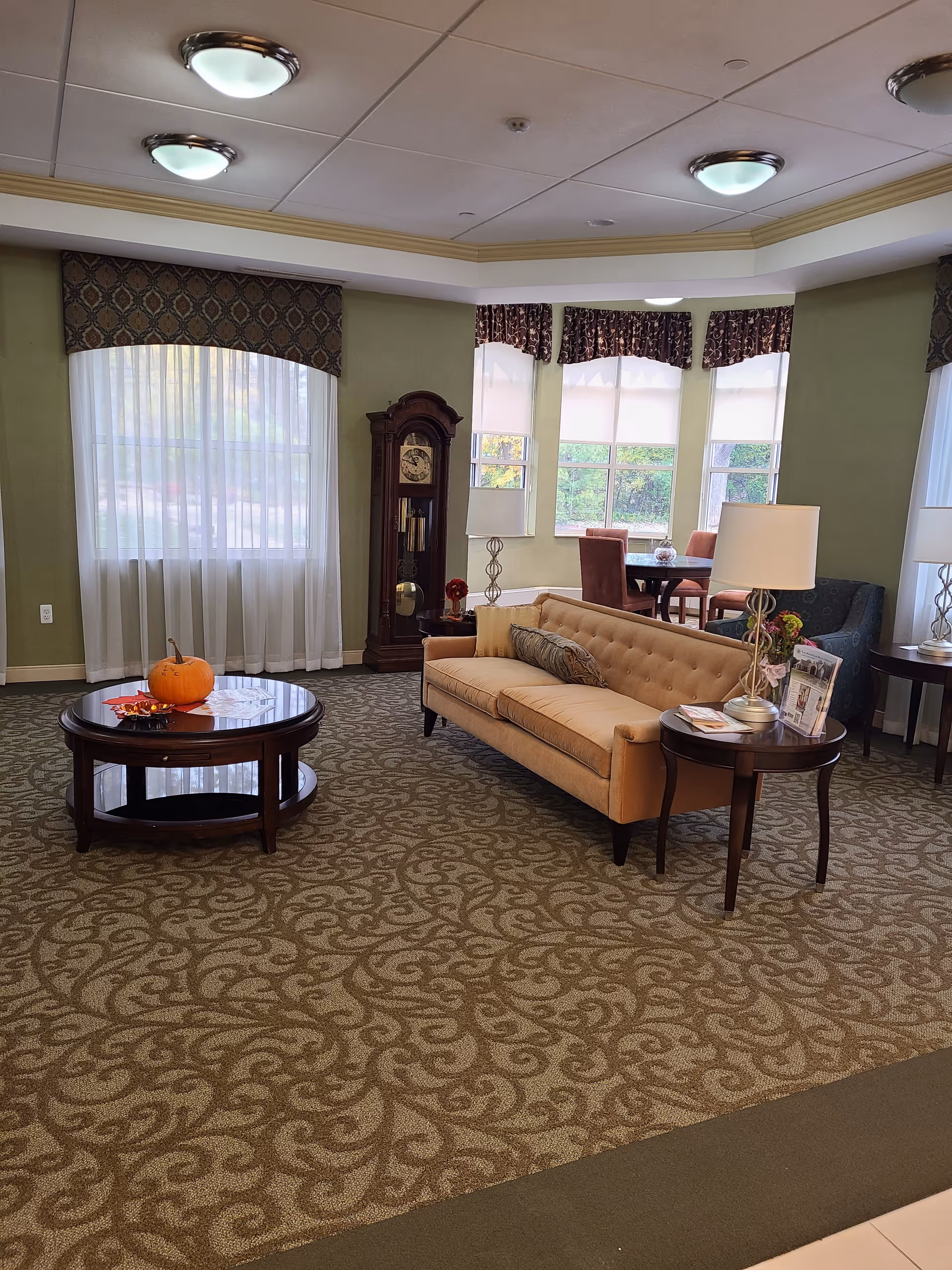 Communal living room with a tufted sofa, round coffee table topped with a pumpkin, side tables and lamps, a grandfather clock, and bay windows with curtains.