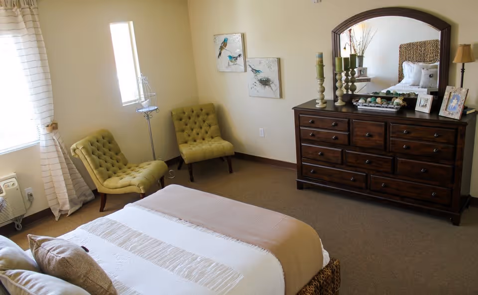 A cozy bedroom with a bed covered in white and beige linens, two green tufted chairs near a window with striped curtains, a dark wooden dresser with a large mirror, decorative candles, framed photos, and bird artwork on the wall.