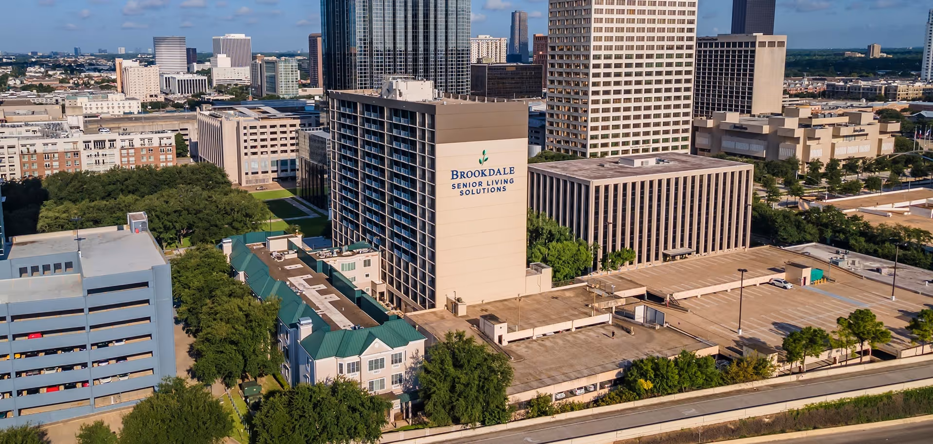 Aerial view of the Brookdale Senior Living Solutions building in an urban area, surrounded by other tall buildings, parking structures, and greenery under a partly cloudy sky.