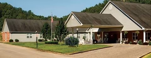Exterior view of a single-story senior living facility building with a covered entrance, well-maintained lawn, shrubs, and an American flag on a flagpole.