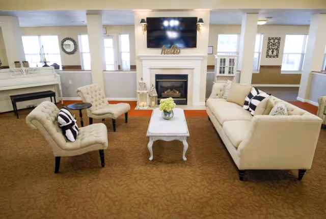 Communal living room with a beige sofa and matching chairs arranged around a white coffee table in front of a fireplace and wall-mounted TV.