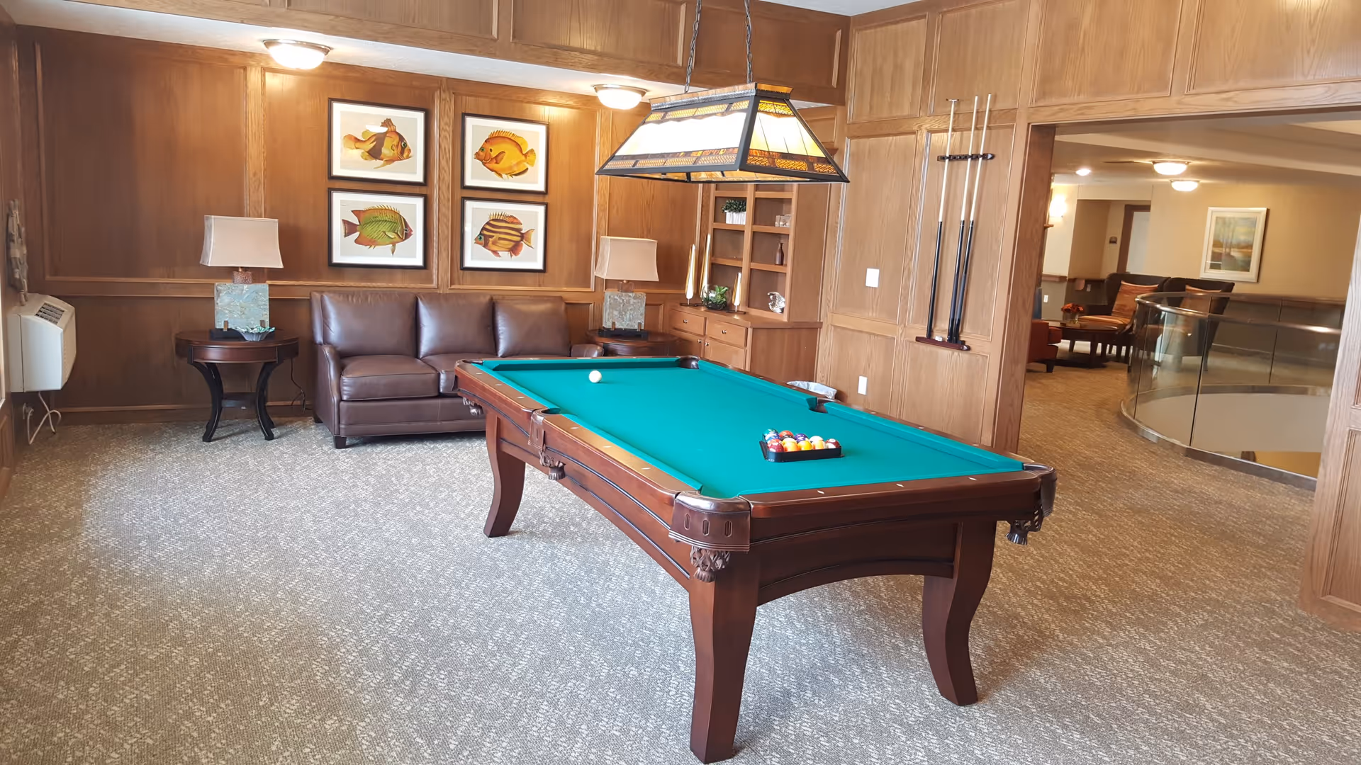 A cozy recreation room with a green felt pool table in the center, a brown leather couch against a wood-paneled wall, two side tables with lamps, and four framed fish illustrations hanging above the couch. The room has beige carpet flooring and a stained glass hanging light fixture above the pool table. In the background, there is an open area with additional seating and a glass railing overlooking a lower level.