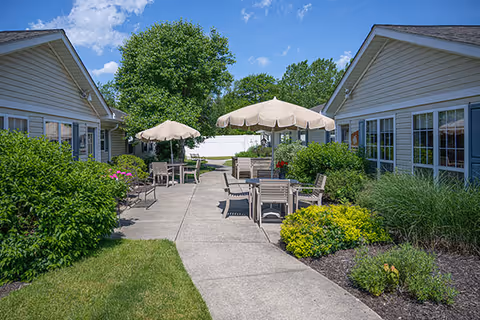 Outdoor patio area between two single-story buildings with beige siding, featuring several tables with umbrellas and chairs surrounded by green bushes and plants under a blue sky with some clouds.