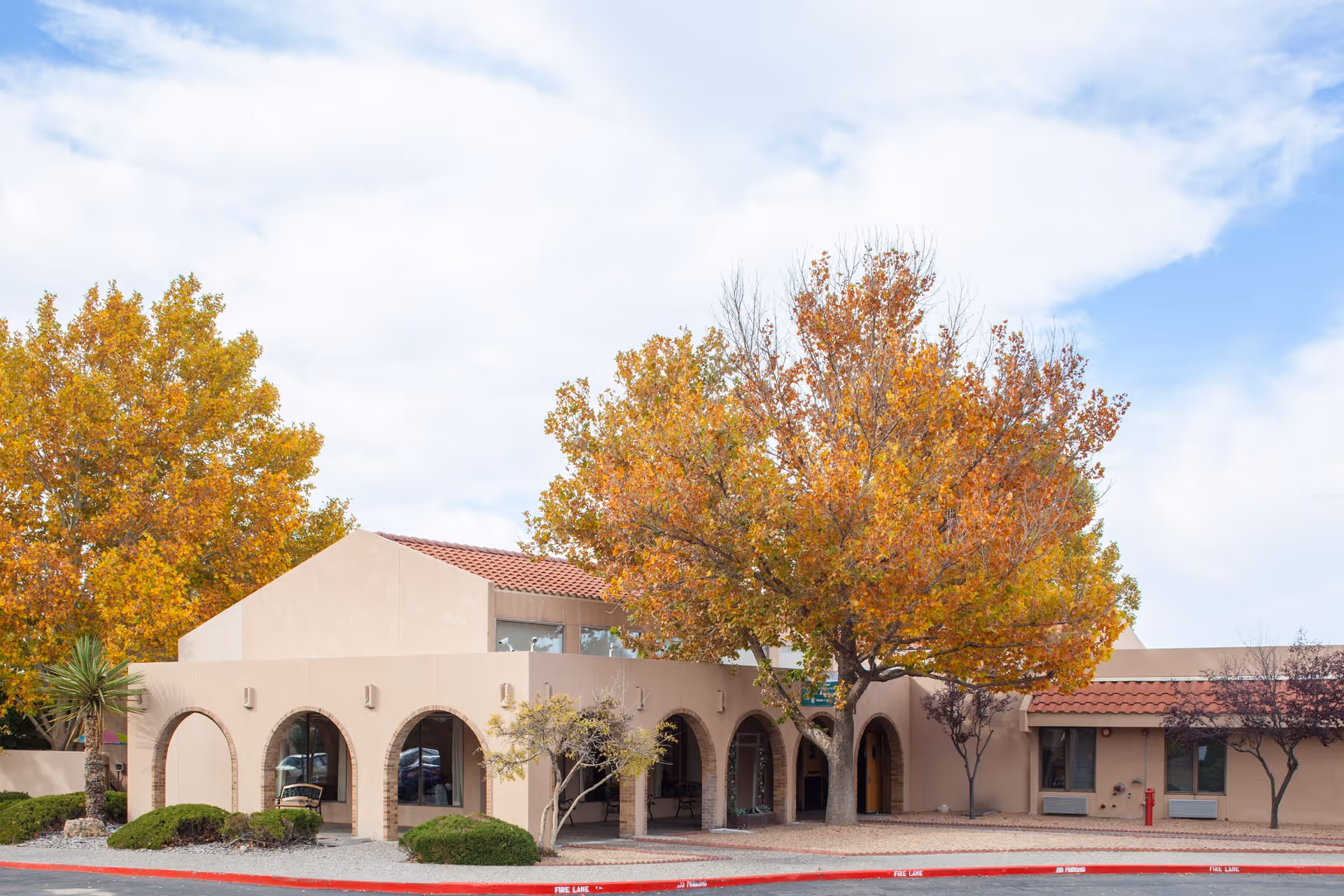 Front exterior of a beige stucco building with arched entrances and large autumn trees in front.
