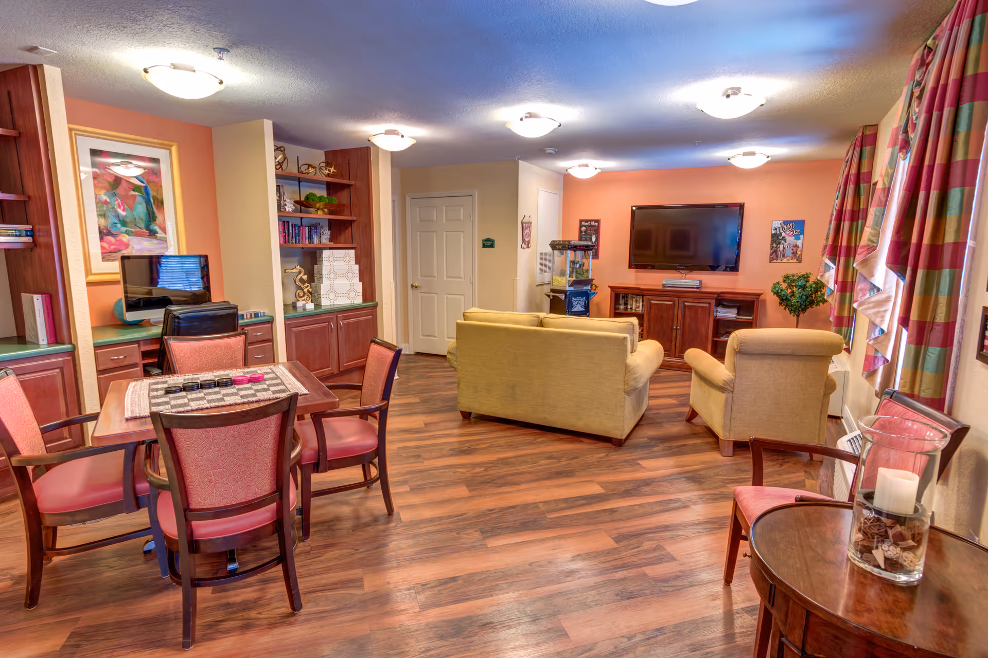Well-lit senior living common room with sofas and armchairs facing a wall-mounted TV, a small dining/game table, bookshelves and wood floors.