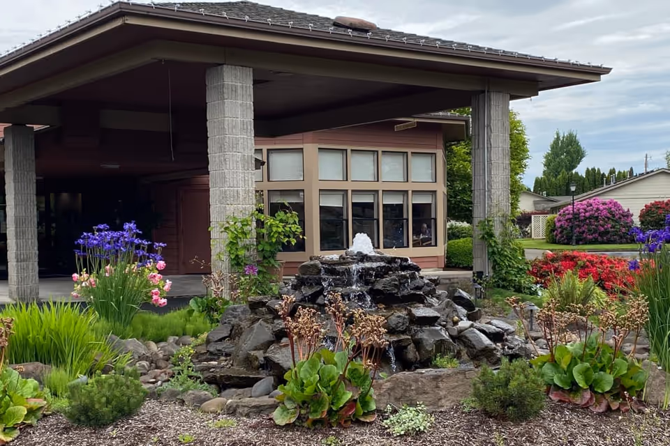 Front entrance of the facility with a covered porte-cochère, a rock fountain and colorful landscaped flower beds.