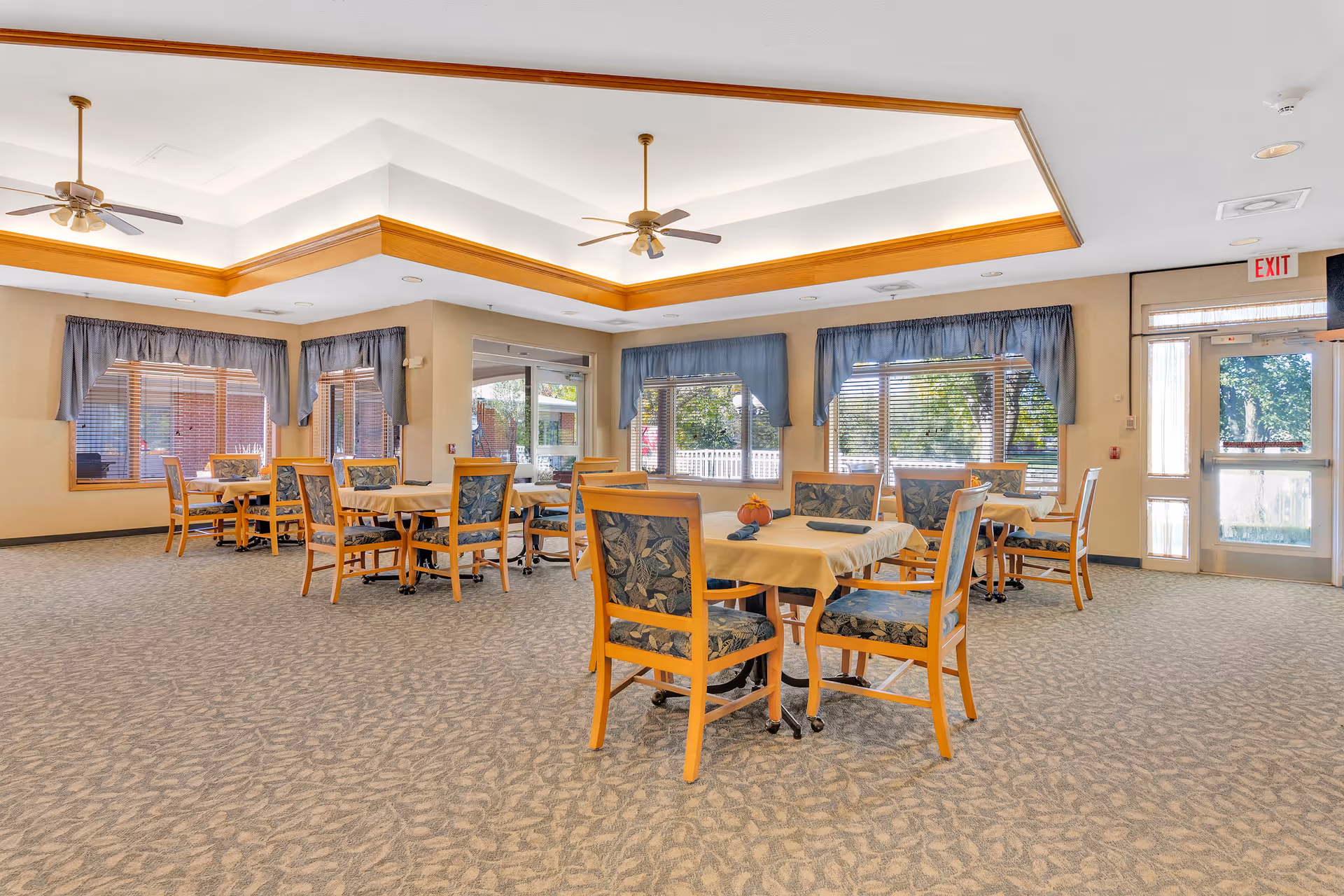 A spacious dining room in a senior living facility with multiple tables covered with beige tablecloths and surrounded by wooden chairs with floral upholstery. The room has large windows with blue curtains allowing natural light to enter, a carpeted floor with a subtle pattern, ceiling fans, and an exit door with a window.