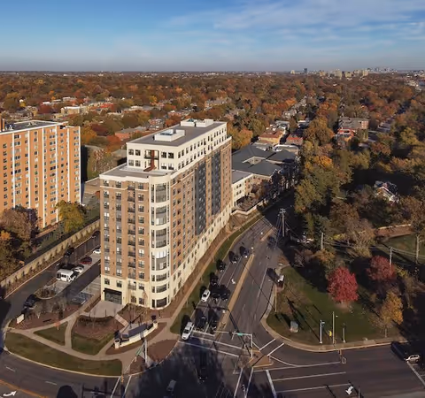 Aerial view of a multi-story residential building surrounded by roads, trees with autumn foliage, and other buildings in the distance under a partly cloudy sky.