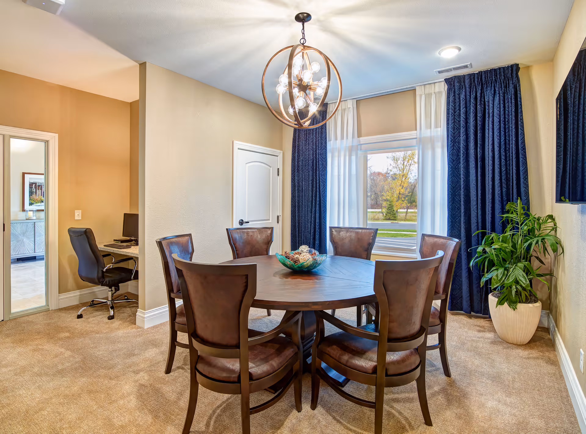 A dining room with a round wooden table surrounded by six brown cushioned chairs. A modern chandelier hangs above the table. There is a large window with sheer white and dark blue curtains letting in natural light. To the left, there is a small office nook with a black office chair and a computer desk. A potted plant is placed near the window on the right side.