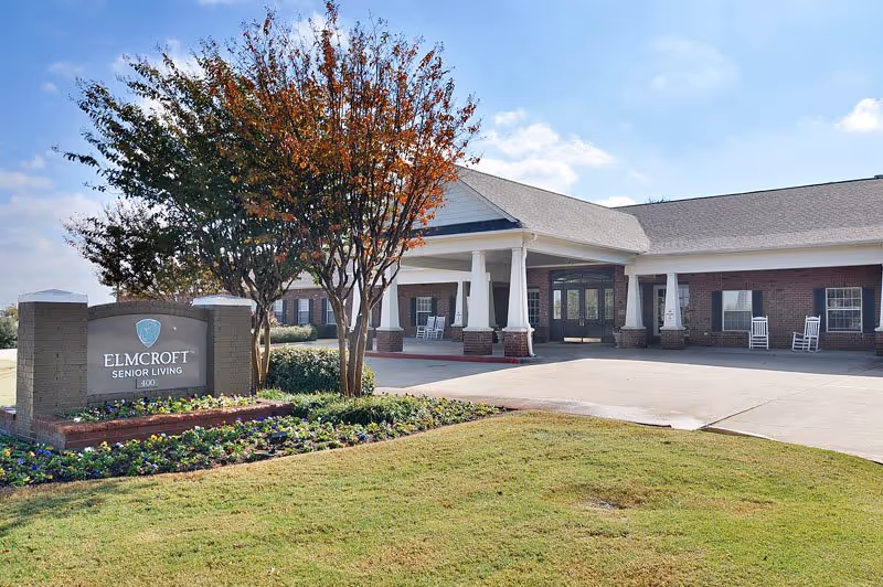 Exterior view of a senior living facility building with a covered entrance, white pillars, and rocking chairs on the porch. In front, there is a landscaped area with a sign that reads 'ELMCROFT SENIOR LIVING'. The sky is clear with some clouds.