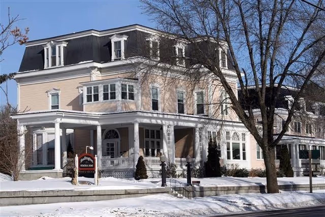Exterior view of a large, three-story historic-style building with a covered porch and multiple windows, surrounded by snow-covered ground and leafless trees under a clear blue sky.