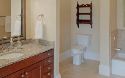 Bathroom with a granite-top vanity and sink, wall mirror and towel, a toilet beneath a decorative wooden shelf, and a partial bathtub visible.