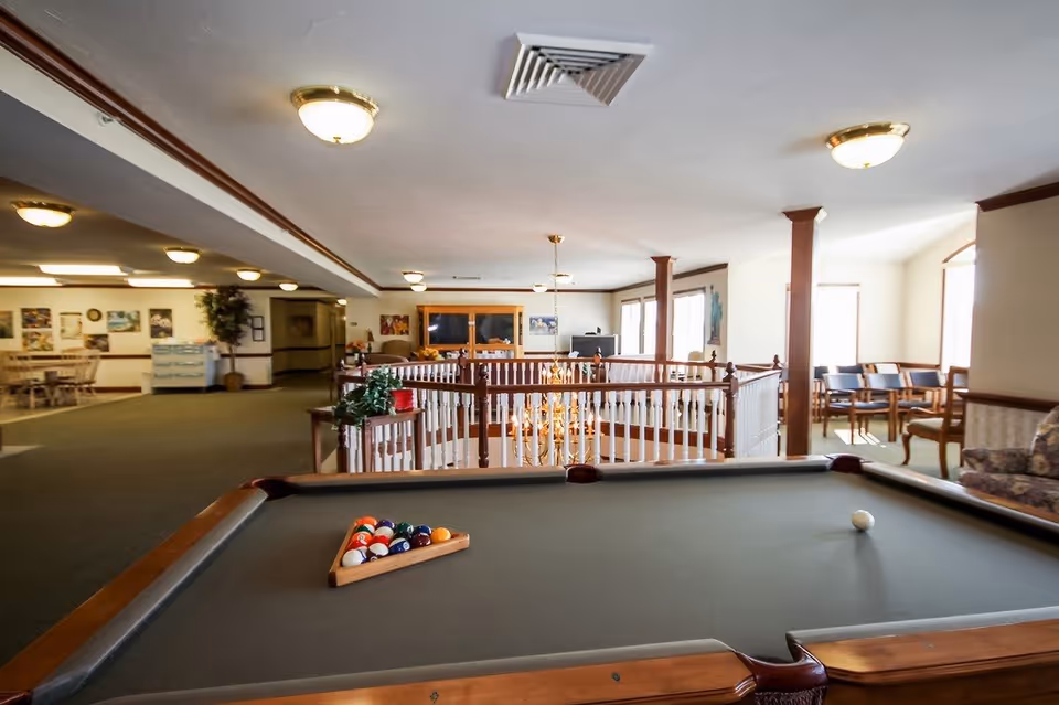 Interior view of a senior living facility common area featuring a pool table in the foreground with billiard balls arranged in a triangle. The room has carpeted floors, multiple seating areas with chairs and sofas, a railing around a stairwell, and several ceiling lights. There are windows letting in natural light and framed pictures on the walls.
