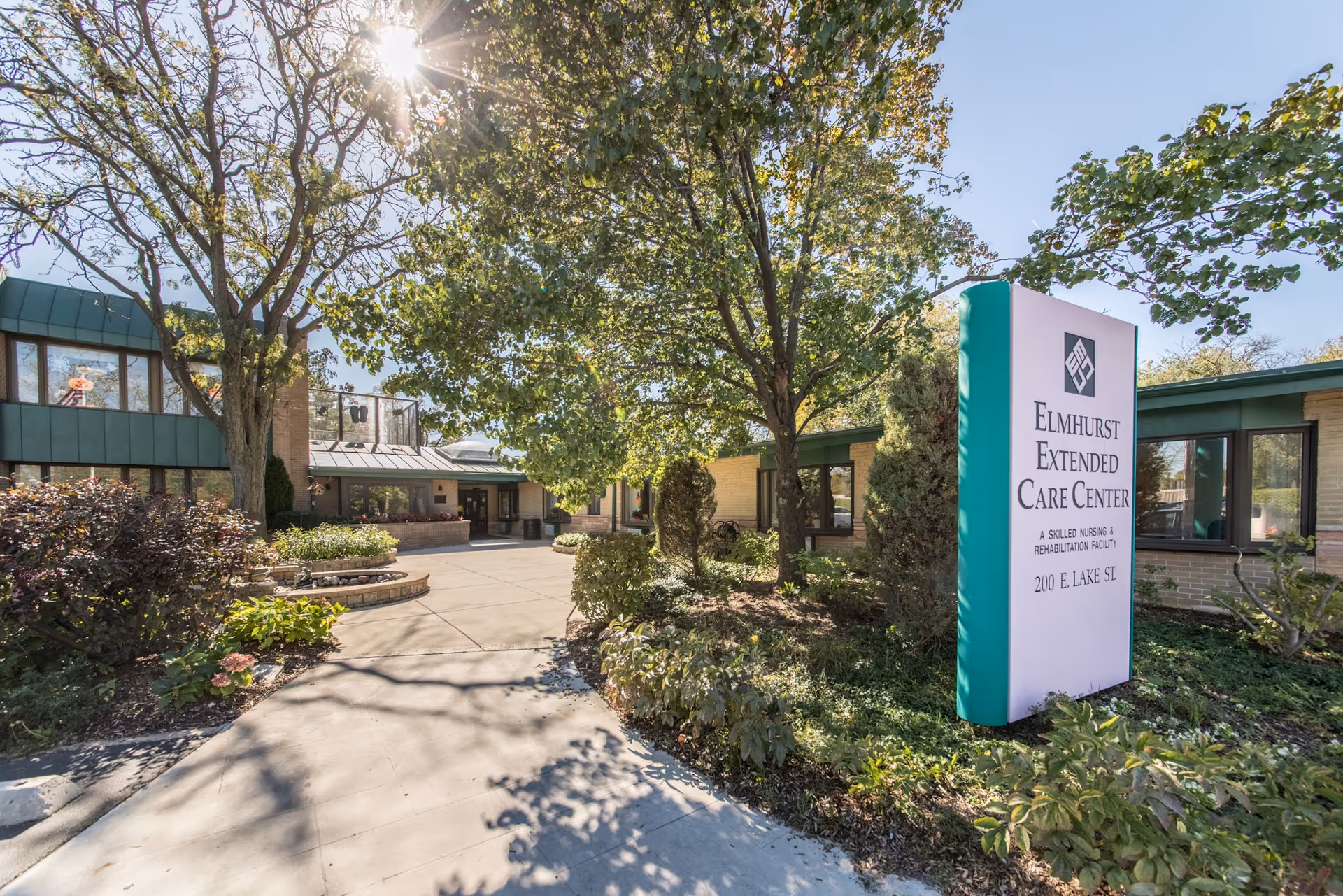 Exterior view of Elmhurst Extended Care Center on a sunny day, showing the building entrance surrounded by trees and shrubs, with a large sign displaying the facility name and address.