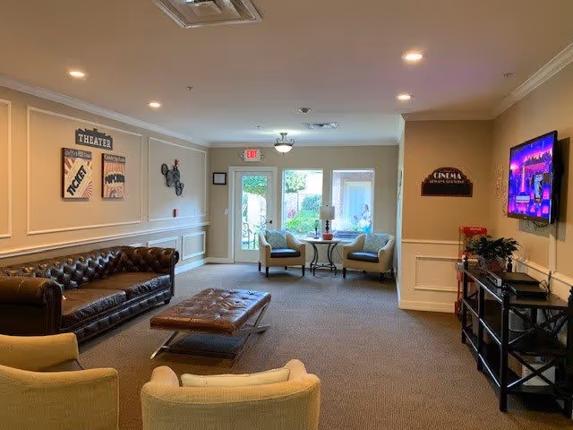 Well-lit common lounge with a leather sofa, armchairs, ottoman, wall-mounted TV and a small seating area by glass doors.