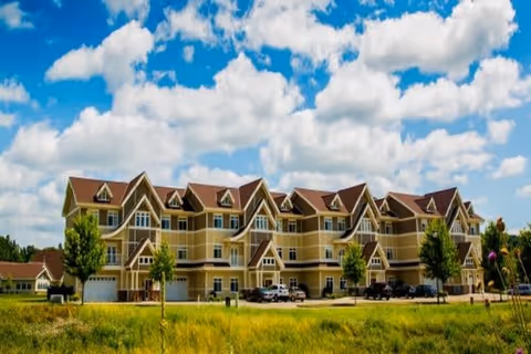 A large, multi-story residential building with beige siding and brown roofs under a partly cloudy blue sky. The building has multiple balconies and windows, with a parking area in front and some small trees and grass in the foreground.