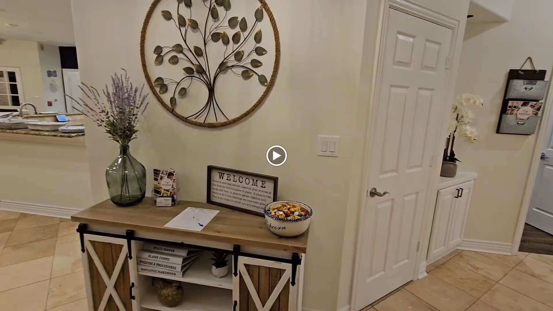 Interior view of a hallway in an assisted living facility featuring a wooden console table with decorative items including a large green vase with flowers, a welcome sign, a bowl of candy, and some paperwork. The walls are light-colored, and there is a decorative metal wall art piece above the table. To the right, there is a closed white door and a small cabinet with a potted orchid on top.