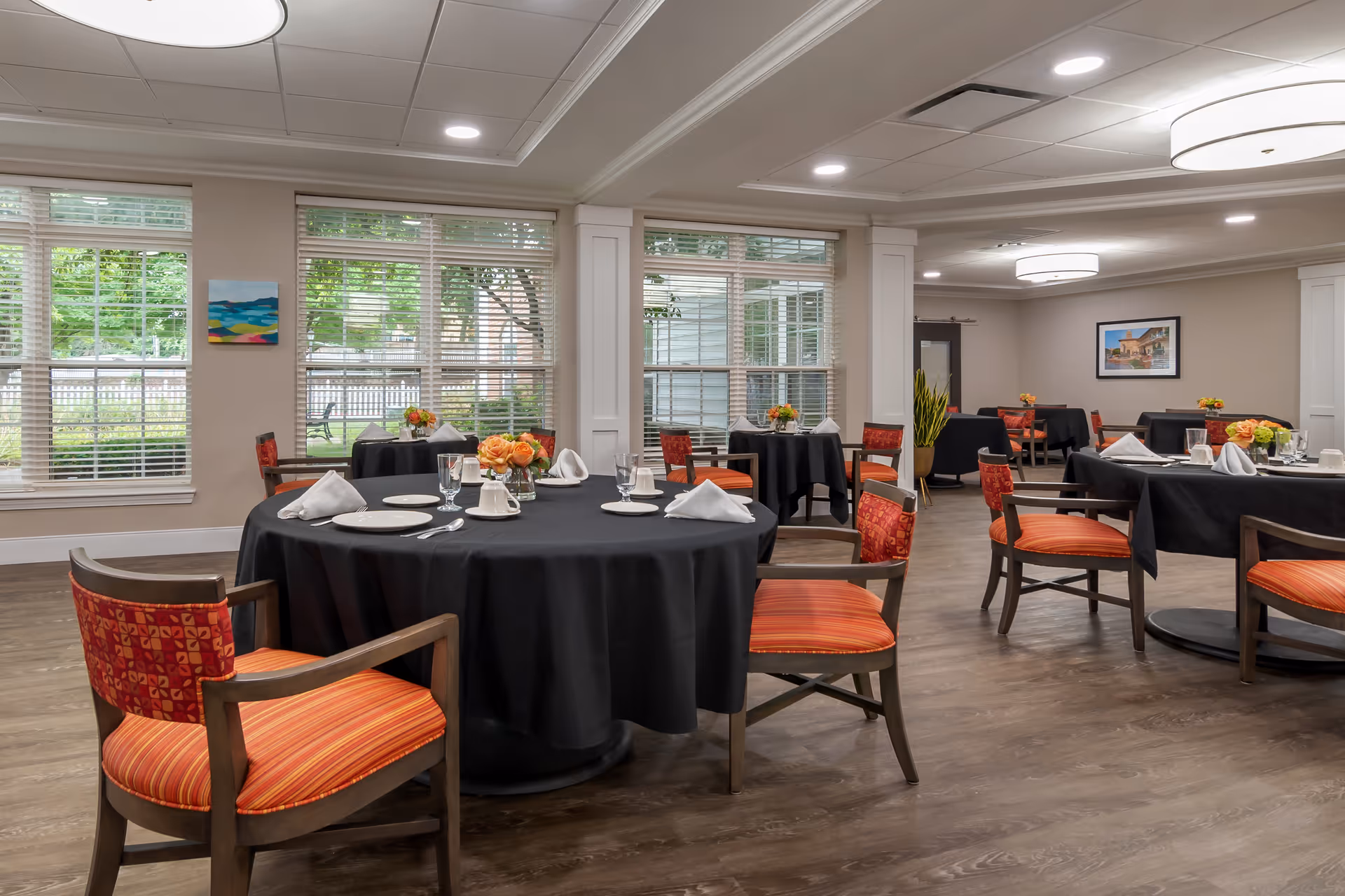 Dining room with round tables covered in black tablecloths, place settings, and wooden chairs with orange cushions in a bright room with large windows.