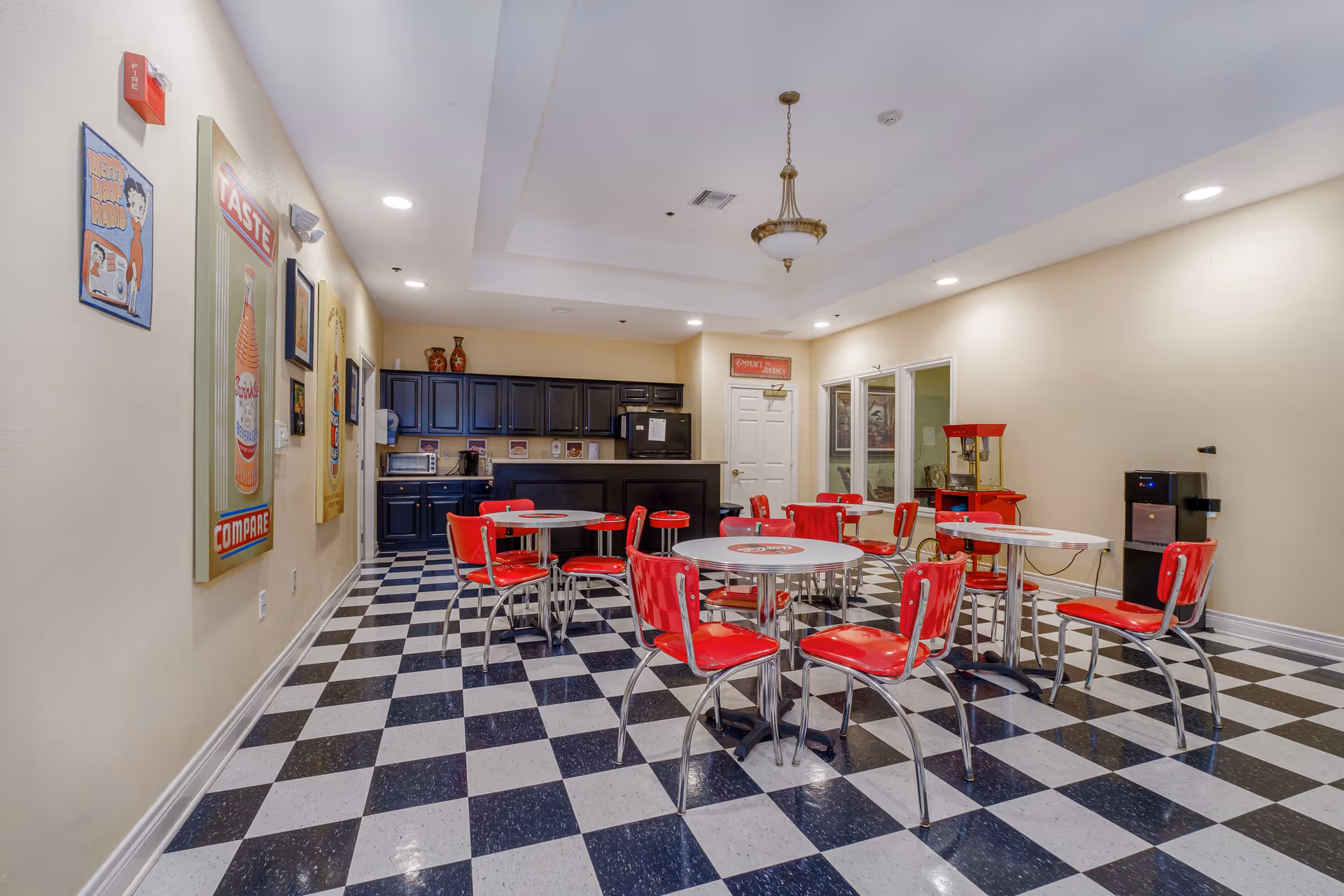 Retro-style communal dining area with red diner chairs and round tables on a black-and-white checkered floor and a kitchenette in the background.