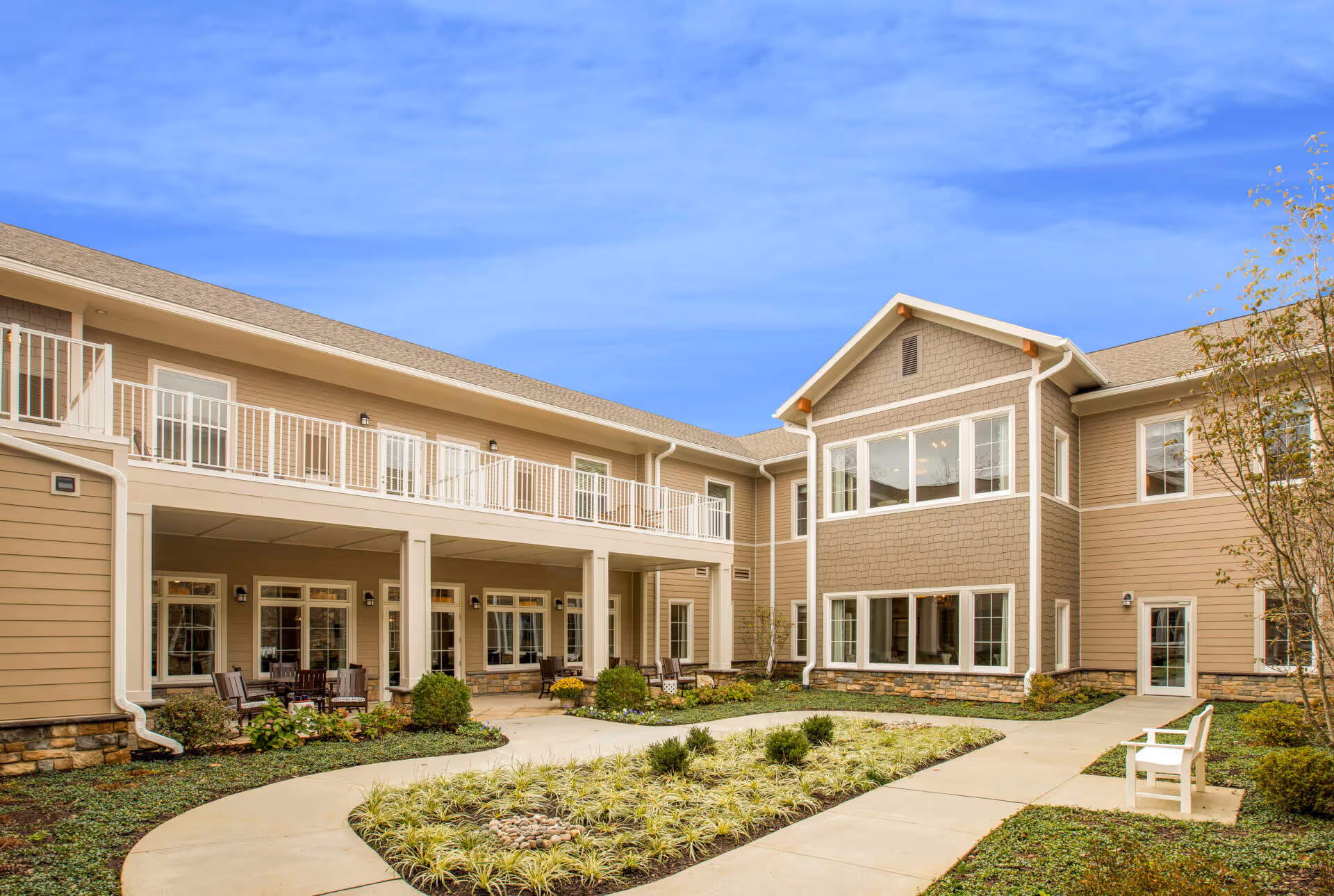 Exterior view of a two-story senior living facility building with beige siding and white trim. The building surrounds a landscaped courtyard with a curved concrete walkway, plants, and a white bench. The sky is clear and blue.