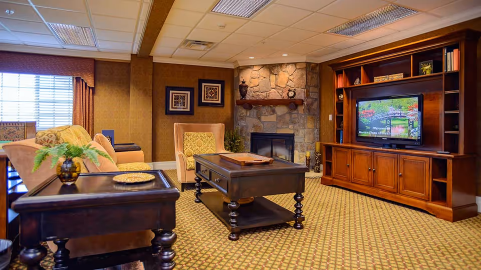 A cozy living room with a stone fireplace, a wooden entertainment center with a TV displaying a welcome message for Garden Plaza Of Greenbriar Cove, a beige sofa, an armchair with a patterned cushion, a wooden coffee table, and a side table with a small plant. The room has patterned carpet and warm lighting.