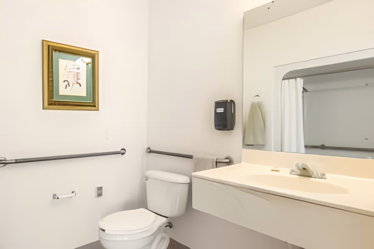 A clean and simple bathroom with a white toilet, a wall-mounted sink with a large mirror above it, a towel hanging on a rail, a soap dispenser mounted on the wall, and a framed picture hanging on the wall.