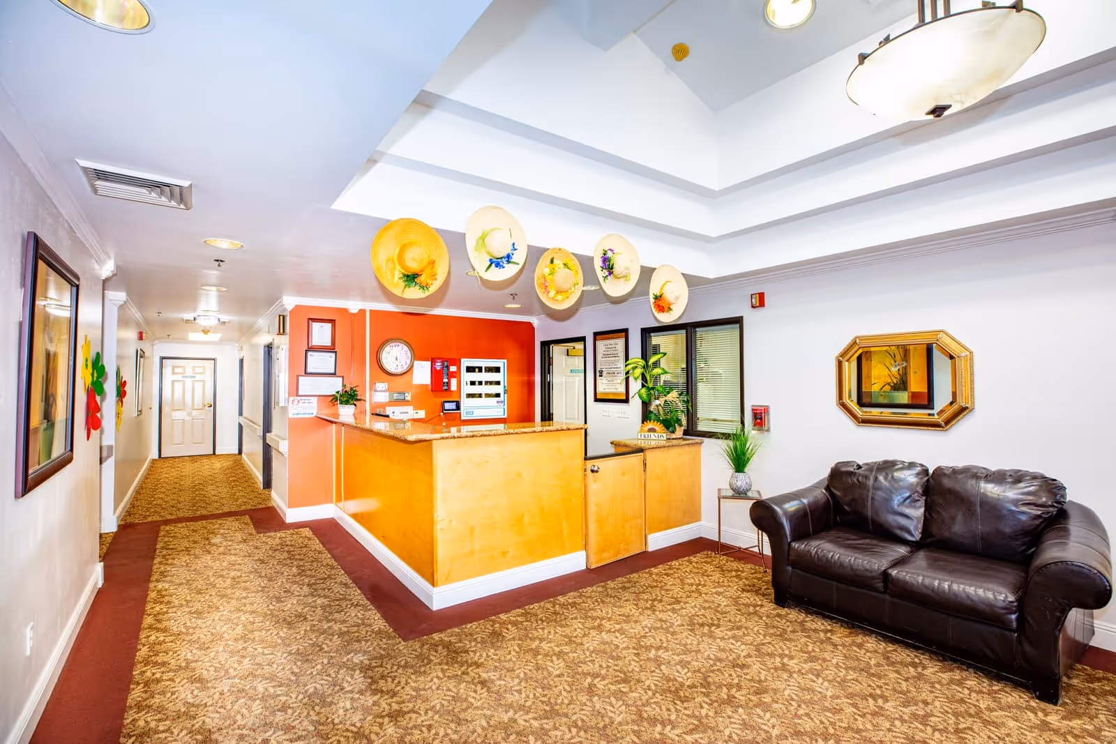 Reception area of Tuscany Villa Senior Living with a wooden front desk, a brown leather couch, a small side table with a plant, decorative hats hanging on the wall above the desk, a mirror on the wall, and a hallway leading to doors in the background.